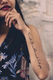 A woman with a tattoo on her forearm featuring cursive writing. She is wearing a dark floral-patterned garment and has dark lipstick. Her hand is near her face, and the background is a light-colored textured wall.