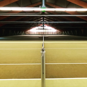 An indoor tennis court is seen from the perspective of the net, with overhead lighting and a roof structure made of wooden beams. The court surface is a muted, earthy color, and no people are present on the court.