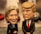 A pair of caricature-style bobblehead dolls depicting two figures with exaggerated facial features. Both are wearing formal attire, one with a wide smile and the other with a serious expression. The background is blurred, focusing attention on the dolls.