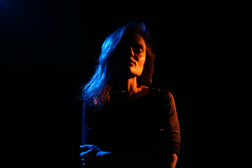 A person stands against a dark background with dramatic lighting casting shadows across their face. The lighting creates a strong contrast with hues of blue and orange gently illuminating parts of their hair and clothing.