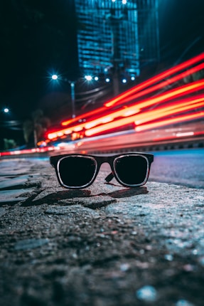 A pair of sunglasses rests on a textured surface in an urban nighttime setting. Bright red light trails from passing vehicles create dynamic lines across the middle of the image, while a tall, illuminated building looms in the background. The scene is lit by streetlights, adding a sense of depth and perspective.