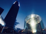 A modern, cylindrical glass structure with a prominent illuminated logo at its center, set against the backdrop of a towering building and a deep blue sky. The towering structure is slightly illuminated, creating a contrast with the dark glass of the cylinder.