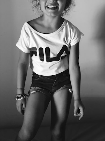 A young person is wearing a casual outfit consisting of a T-shirt with 'FILA' written on it and denim shorts. They are smiling, and their joyful expression complements the relaxed pose against a plain background.