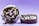 Two chocolate donuts with white icing and dark cookie crumb toppings are displayed against a gradient purple background. One donut is upright, leaning slightly on the other, while the second donut lies flat. Crumbs are scattered around the base, enhancing the sense of texture.