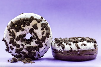 Two chocolate donuts with white icing and dark cookie crumb toppings are displayed against a gradient purple background. One donut is upright, leaning slightly on the other, while the second donut lies flat. Crumbs are scattered around the base, enhancing the sense of texture.