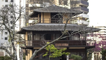A traditional pagoda-style building is nestled among urban structures, surrounded by leafless trees with a few green leaves. The architectural design showcases wooden railings and a tiered roof, creating a serene contrast against the backdrop of modern high-rise buildings.