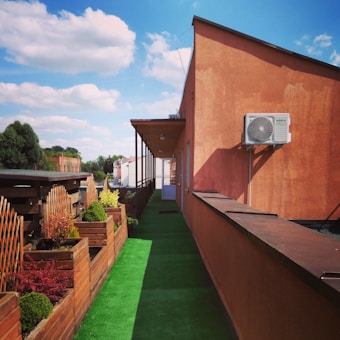 A rooftop area with a green artificial grass pathway flanked by wooden planters filled with various plants. An orange building wall with an air conditioning unit is prominently visible. The sky is clear with a few fluffy clouds, adding a serene backdrop.