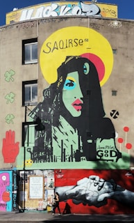 A vibrant mural features a colorful portrait of a woman with stylized facial features. The artwork includes a wide yellow halo with the word 'SAOIRSE' above the woman's head. Surrounding elements include a red hand, clover, and abstract symbols. The lower part of the mural depicts two hands clasping against a red background. Various graffiti and tags adorn the upper and lower sections of the building.