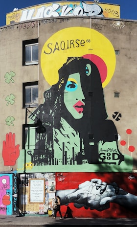 A vibrant mural features a colorful portrait of a woman with stylized facial features. The artwork includes a wide yellow halo with the word 'SAOIRSE' above the woman's head. Surrounding elements include a red hand, clover, and abstract symbols. The lower part of the mural depicts two hands clasping against a red background. Various graffiti and tags adorn the upper and lower sections of the building.