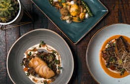 Three aesthetically plated dishes are arranged on a rustic wooden table. The dish on the bottom left features a piece of cooked fish surrounded by various garnishes and sauces on a gray plate. The upper dish is a vibrant vegetarian display with yellow squash, greens, and decorative flowers on a teal rectangular plate. The bottom right dish consists of a piece of seasoned meat accompanied by beans and sprouts, presented on a white plate with an orange sauce.