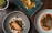 Three aesthetically plated dishes are arranged on a rustic wooden table. The dish on the bottom left features a piece of cooked fish surrounded by various garnishes and sauces on a gray plate. The upper dish is a vibrant vegetarian display with yellow squash, greens, and decorative flowers on a teal rectangular plate. The bottom right dish consists of a piece of seasoned meat accompanied by beans and sprouts, presented on a white plate with an orange sauce.