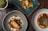 Three aesthetically plated dishes are arranged on a rustic wooden table. The dish on the bottom left features a piece of cooked fish surrounded by various garnishes and sauces on a gray plate. The upper dish is a vibrant vegetarian display with yellow squash, greens, and decorative flowers on a teal rectangular plate. The bottom right dish consists of a piece of seasoned meat accompanied by beans and sprouts, presented on a white plate with an orange sauce.
