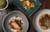 Three aesthetically plated dishes are arranged on a rustic wooden table. The dish on the bottom left features a piece of cooked fish surrounded by various garnishes and sauces on a gray plate. The upper dish is a vibrant vegetarian display with yellow squash, greens, and decorative flowers on a teal rectangular plate. The bottom right dish consists of a piece of seasoned meat accompanied by beans and sprouts, presented on a white plate with an orange sauce.