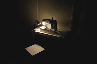 A sewing machine is set up on a tidy desk, illuminated by a small, focused desk lamp. The surrounding area is dark, emphasizing the spotlight effect on the sewing machine and workspace. The atmosphere appears serene and concentrated, suitable for creating or working late into the night.