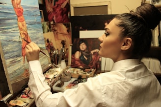 A focused artist paints on a canvas depicting a woman at the beach. The workspace is filled with brushes, paint tubes, and other artworks, indicating a rich, creative environment. The artist is wearing a white shirt and has their hair tied up in a bun, deeply engrossed in their work.