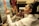 A focused artist paints on a canvas depicting a woman at the beach. The workspace is filled with brushes, paint tubes, and other artworks, indicating a rich, creative environment. The artist is wearing a white shirt and has their hair tied up in a bun, deeply engrossed in their work.