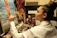 A focused artist paints on a canvas depicting a woman at the beach. The workspace is filled with brushes, paint tubes, and other artworks, indicating a rich, creative environment. The artist is wearing a white shirt and has their hair tied up in a bun, deeply engrossed in their work.