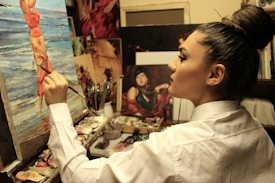 A focused artist paints on a canvas depicting a woman at the beach. The workspace is filled with brushes, paint tubes, and other artworks, indicating a rich, creative environment. The artist is wearing a white shirt and has their hair tied up in a bun, deeply engrossed in their work.