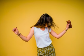 A person with long hair is shown in mid-motion against a vibrant yellow background. They are wearing a white sleeveless top and green patterned pants, holding a small brown object in their right hand. Their hair swings across their face as they appear to be dancing.