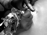 A person wearing black gloves is using a tattoo machine to apply a design on skin. The image focuses on the tattoo machine and the hand holding it, emphasizing the intricacy of the tattooing process.