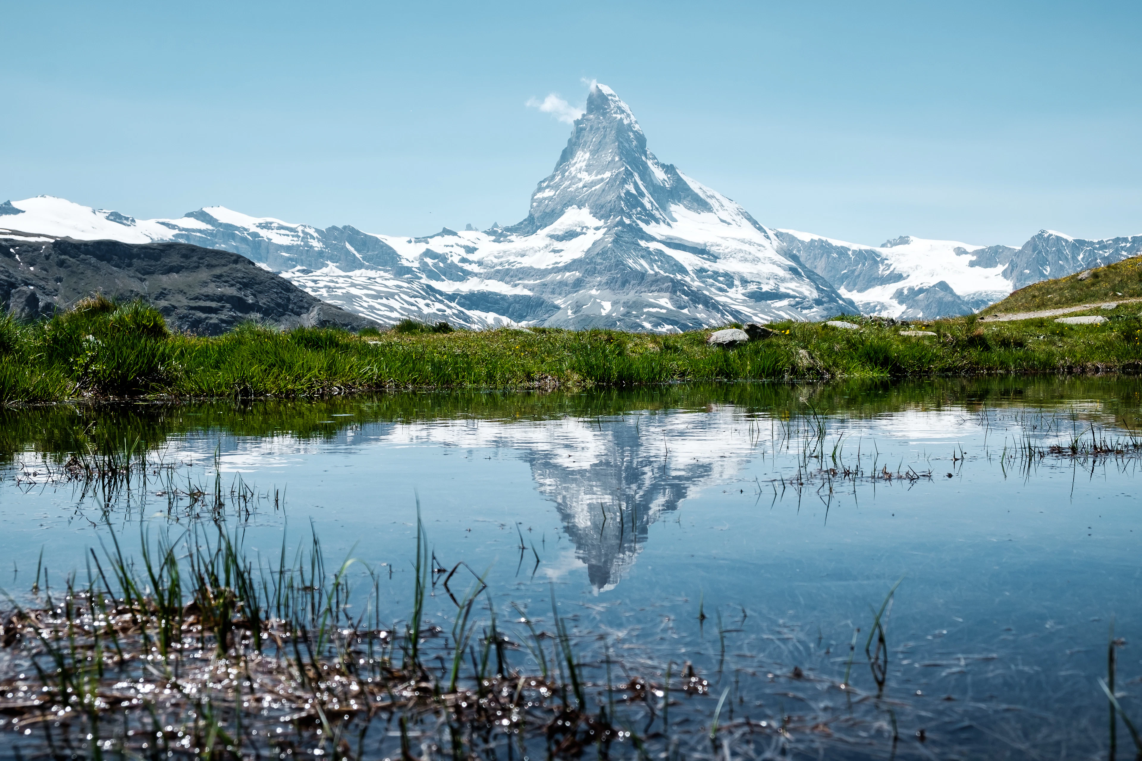 The snow-capped Matterhorn and its reflection mirror perfectly in a still alpine lake fringed with grass.