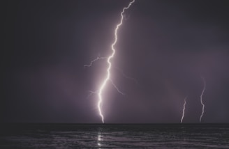 A dramatic scene of lightning striking the ocean, with two visible bolts illuminating the dark, cloudy sky. The surface of the water reflects the bright, jagged lines of the lightning.