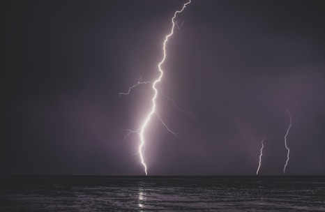 A dramatic scene of lightning striking the ocean, with two visible bolts illuminating the dark, cloudy sky. The surface of the water reflects the bright, jagged lines of the lightning.