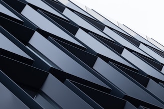 A series of overlapping angular metal panels create a geometric pattern on a building facade. The panels are aligned in a repetitive, diagonal arrangement that gives a sense of depth and modern architectural design.