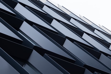 A series of overlapping angular metal panels create a geometric pattern on a building facade. The panels are aligned in a repetitive, diagonal arrangement that gives a sense of depth and modern architectural design.