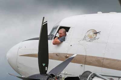 A man leans out of the cockpit window of an airplane, which is marked with the name 'Alexandra' and a logo featuring a globe and airplane. The propeller is visible in the foreground and the sky is overcast.