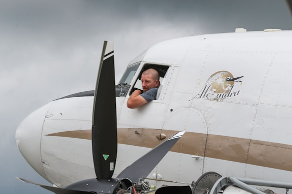 A man leans out of the cockpit window of an airplane, which is marked with the name 'Alexandra' and a logo featuring a globe and airplane. The propeller is visible in the foreground and the sky is overcast.