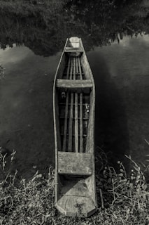 A wooden canoe is positioned in still water, surrounded by partially visible foliage. The perspective is from above, highlighting the canoe's rustic, weathered texture as it floats peacefully. The water's surface reflects the surrounding greenery and sky, creating a serene and natural atmosphere.