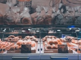 A well-lit bakery display featuring a variety of baked goods, including loaves of bread and pastries arranged in wooden trays inside a glass case. Above the display, several bags of bread are neatly lined up. The background includes a wall with a mural depicting bread and bakery themes.