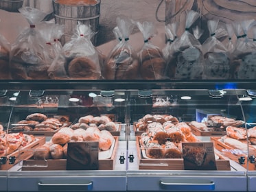 A well-lit bakery display featuring a variety of baked goods, including loaves of bread and pastries arranged in wooden trays inside a glass case. Above the display, several bags of bread are neatly lined up. The background includes a wall with a mural depicting bread and bakery themes.