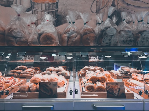 A well-lit bakery display featuring a variety of baked goods, including loaves of bread and pastries arranged in wooden trays inside a glass case. Above the display, several bags of bread are neatly lined up. The background includes a wall with a mural depicting bread and bakery themes.