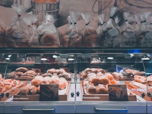 A well-lit bakery display featuring a variety of baked goods, including loaves of bread and pastries arranged in wooden trays inside a glass case. Above the display, several bags of bread are neatly lined up. The background includes a wall with a mural depicting bread and bakery themes.