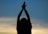 A silhouette of a person with arms raised against a sky that transitions from blue to light orange, creating a serene and contemplative scene. The person appears to be holding their hands together, potentially in a gesture of meditation or prayer.