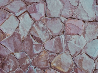 An irregular pattern of overlapping stone tiles, each varying in shape and size, with muted tones of pale pink, grey, and beige. The stones have a rough texture and some gaps are visible between them.