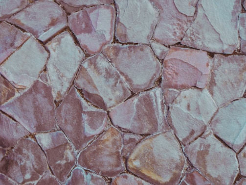 An irregular pattern of overlapping stone tiles, each varying in shape and size, with muted tones of pale pink, grey, and beige. The stones have a rough texture and some gaps are visible between them.