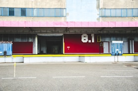 A loading dock area of an industrial building, featuring a red facade with the number 8.1 in white. It includes metal doors and loading ramps, with a pink roof trim and several windows above the dock.