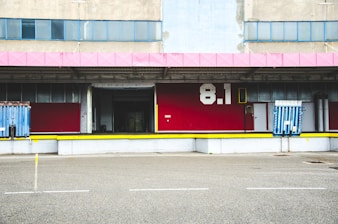 A loading dock area of an industrial building, featuring a red facade with the number 8.1 in white. It includes metal doors and loading ramps, with a pink roof trim and several windows above the dock.