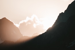 The image features a breathtaking mountain landscape with sharp, dark silhouettes of peaks against a bright sky. Sunlight peeks through between the peaks, casting a warm glow and creating soft highlights and shadows. Clouds float gently above the mountains, adding a sense of tranquility and vastness.
