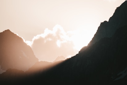 The image features a breathtaking mountain landscape with sharp, dark silhouettes of peaks against a bright sky. Sunlight peeks through between the peaks, casting a warm glow and creating soft highlights and shadows. Clouds float gently above the mountains, adding a sense of tranquility and vastness.