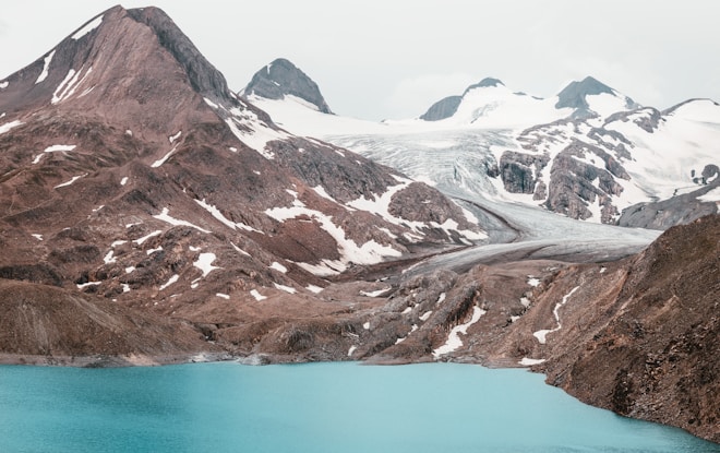 Snow-capped mountains rise majestically, with a glacier flowing down into a vivid turquoise lake. The landscape is rugged with patches of snow nestled against the rocky slopes.