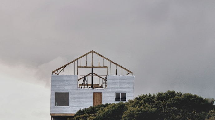 An incomplete wooden frame of a house sits atop a finished lower section with white siding. The structure is set against a overcast sky, and part of it is obscured by dense green foliage at the bottom of the image.