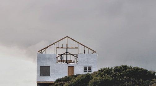 An incomplete wooden frame of a house sits atop a finished lower section with white siding. The structure is set against a overcast sky, and part of it is obscured by dense green foliage at the bottom of the image.