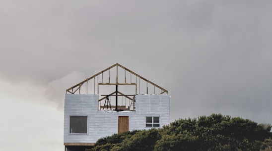 An incomplete wooden frame of a house sits atop a finished lower section with white siding. The structure is set against a overcast sky, and part of it is obscured by dense green foliage at the bottom of the image.
