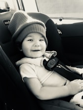 A young child with a playful smile is seated in a car seat, wearing a cozy, oversized knit hat. The setting is inside a vehicle with visible seatbelt fasteners and upholstery. The image has a warm, candid feel.