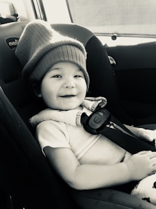 A young child with a playful smile is seated in a car seat, wearing a cozy, oversized knit hat. The setting is inside a vehicle with visible seatbelt fasteners and upholstery. The image has a warm, candid feel.