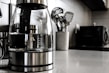 A modern kitchen countertop features a glass electric kettle with a stainless steel base, filled partially with water. Nearby, a white container holds several metallic cooking utensils such as spatulas and ladles. In the background, a microwave oven and other kitchen appliances are slightly blurred.